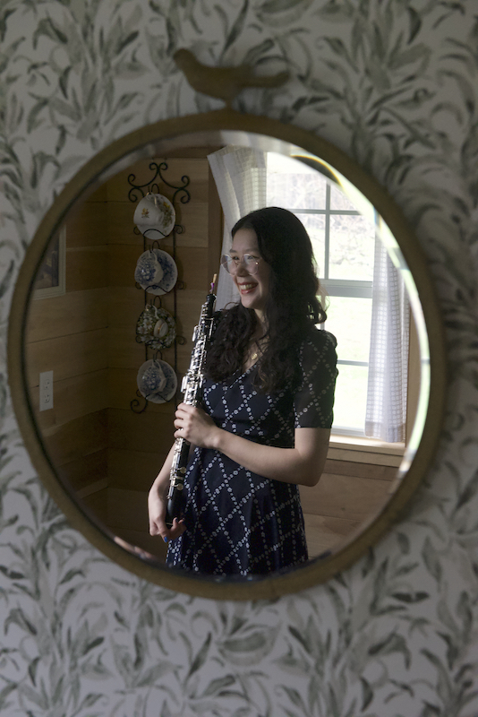 Kiara Hosie, a 25 year old Chinese-Canadian woman
          with long dark hair and glasses, holds an oboe and smiles.
